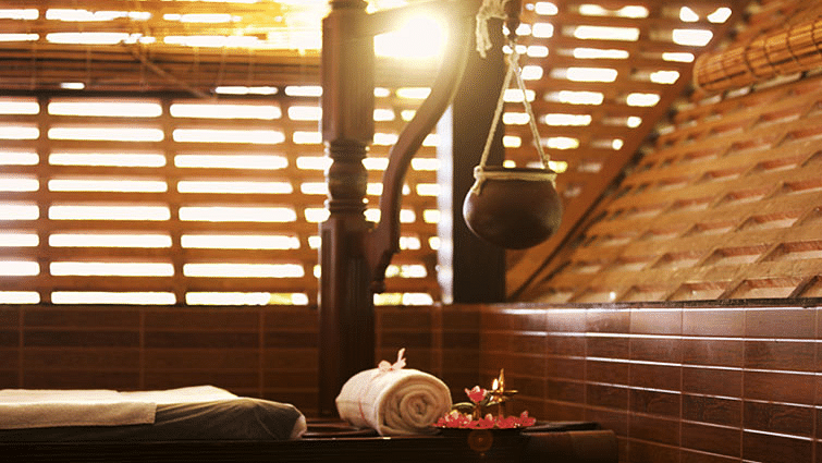 A pot hanging from a wooden bar over the spa bed next to which is a rolled-in towel and flowers at Ayur Ashram, Uday Suites