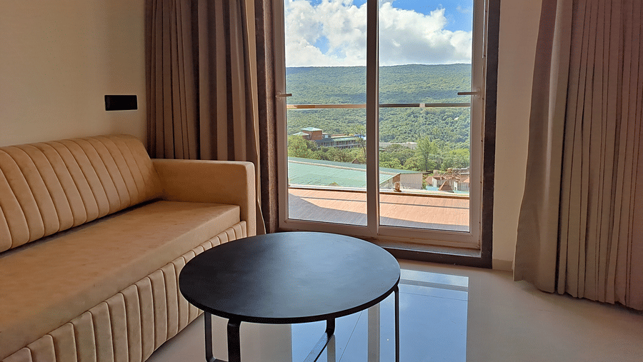 Room with a cushioned sofa, round table, and balcony overlooking green hills