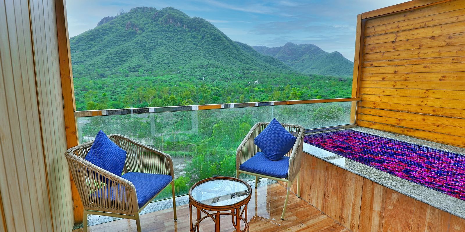A balcony with two chairs, cushions, and a table overlooks a lush green valley with mountains as a scenery.- Bamboo Saa Resort & Spa, Udaipur