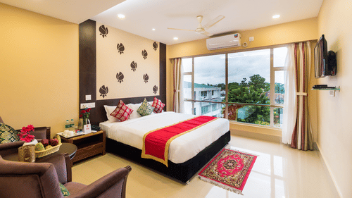A bedroom featuring a neatly arranged bed, a bedside table, sofas on the side, and a balcony with glass windows