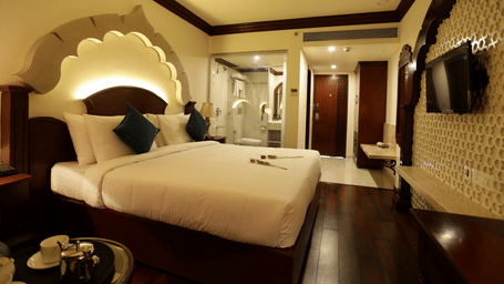 A bedroom at Comfort Inn Sapphire by Eastlynn Collections, Jaipur with queen size bed, arch headboard, side table, wall-mouted television, and view of room entrance.