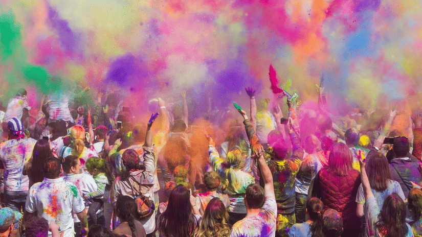A large crowd celebrating outdoors while throwing coloured powder into the air during a festival gathering.