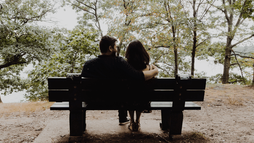 A silhouette of two people sitting on a bench with trees in the background.