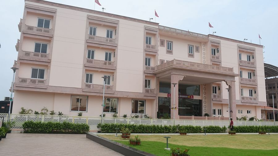 The front view of a modern, light-coloured hotel building with a covered entrance and green lawns in front is presented - Kankidham Lords Inn
