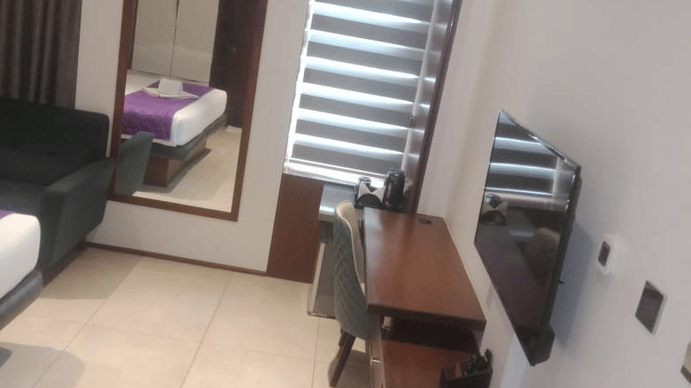 A slightly tilted view of the expty floor space of the Executive Suite at SRS Suites, Koramangala, featuring a wall-mounted television and a window covered with blinds at the end.