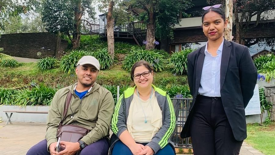 Three people, two seated and one standing, smile for a photo on a wooden bench in a garden setting with lush greenery, trees, and a charming resort building in the background.