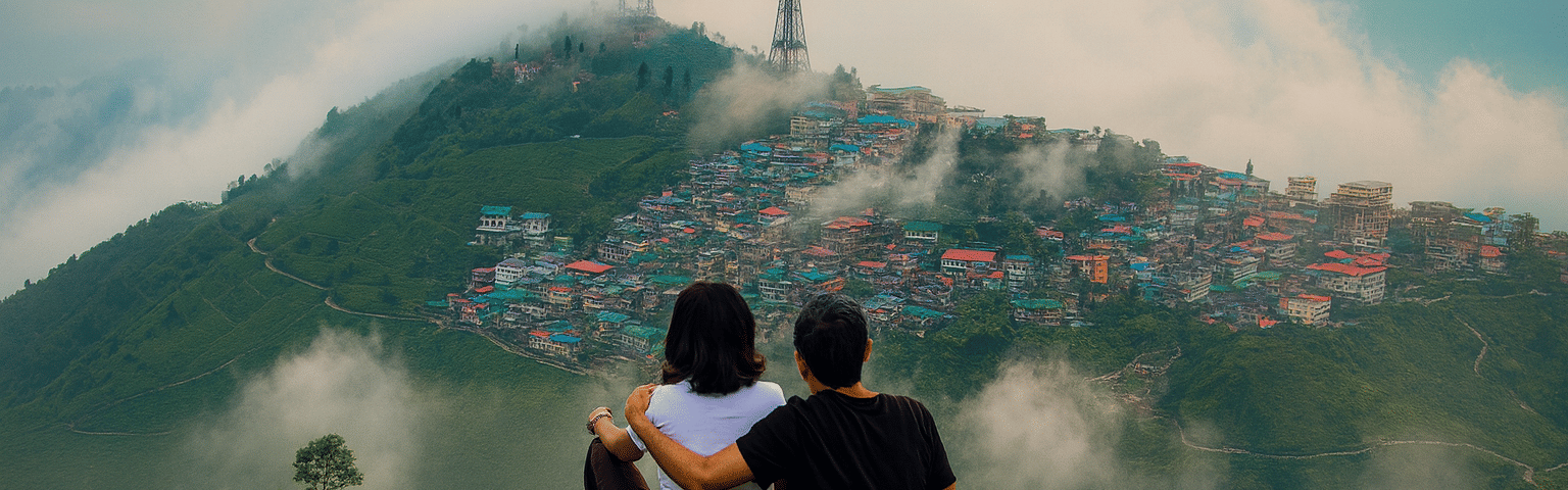 A couple sitting on a hilltop overlooking the misty mountains and colorful cityscape of Kurseong, with clouds floating over the valley, creating a premium and aesthetic morning vibe.