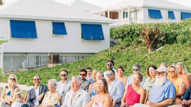 Wedding guests sit in rows of chairs on grass outside blue and white buildings at Abaco Inn.