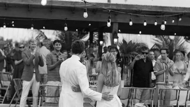 Couple holds hands facing each other on green grass while guests stand nearby, palm tree and shaded area in background at Abaco Inn.