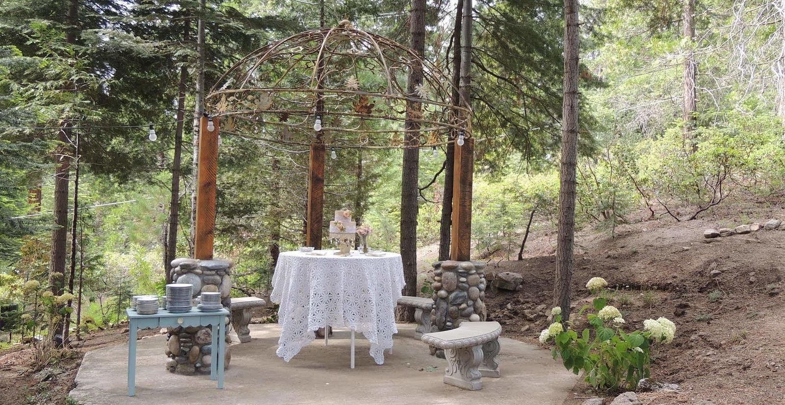 A wedding cake placed on a table in the middle of a forest with a stack of plates placed on a table beside it