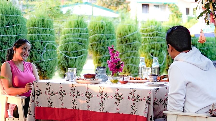 A corner view of a dining area where a couple is gazing at each other, with a table covered in a tablecloth, decorated with a flower vase, food, and a water bottle, all surrounded by plants and trees.