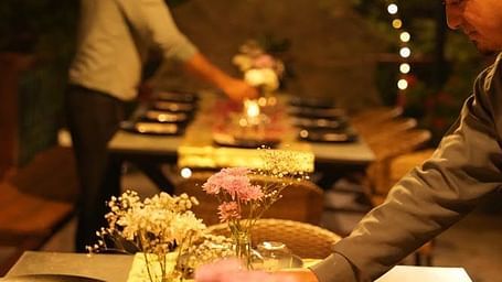Dining table at Karma Chalets set with plates and a flower garland, with a person arranging the setup under lights.