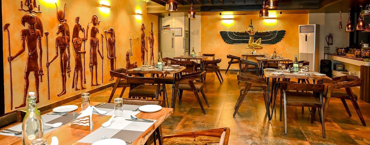 A detailed view of a dining table set for guests in the restaurant, with a focus on the wooden furniture and the warm, ambient lighting from the ceiling fixtures - Sea Hills Hotels, Sri Vijaya Puram (Port Blair)