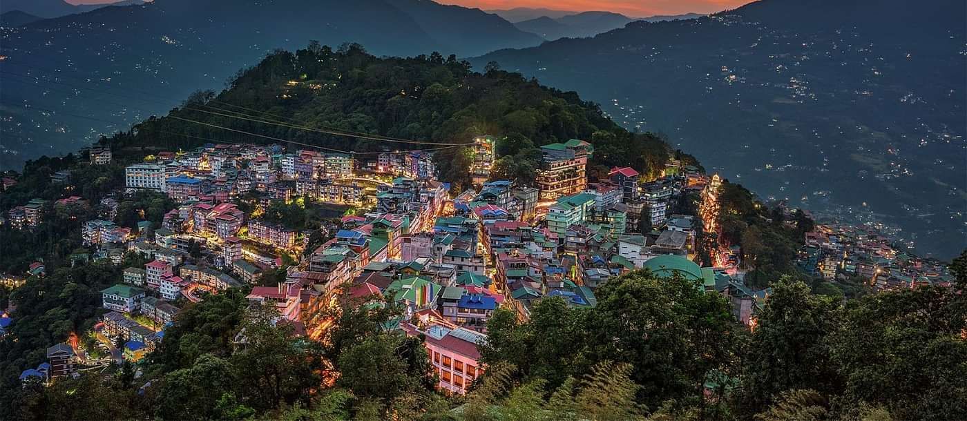 an Ariel view of a city in the hilly regions in the evening with an orange sky with mountain peaks in the background 