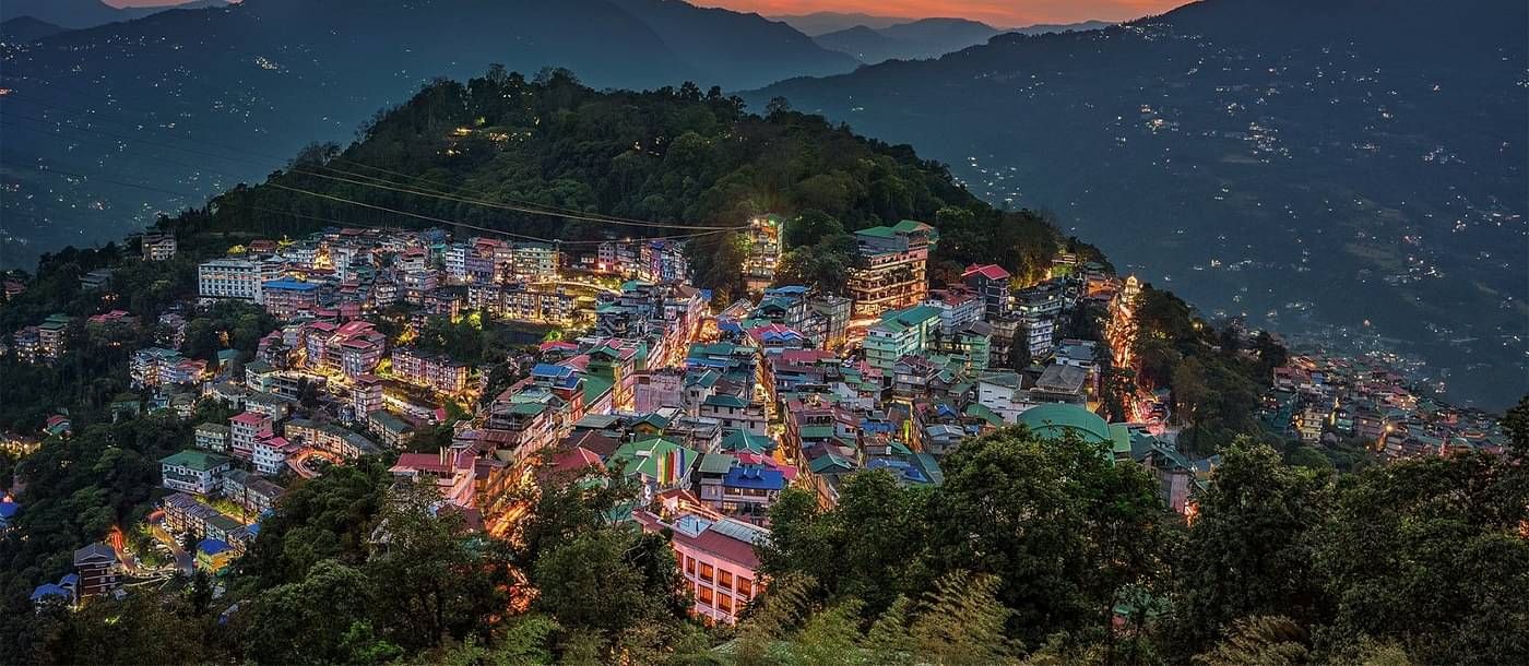 an Ariel view of a city in the hilly regions in the evening with an orange sky with mountain peaks in the background 