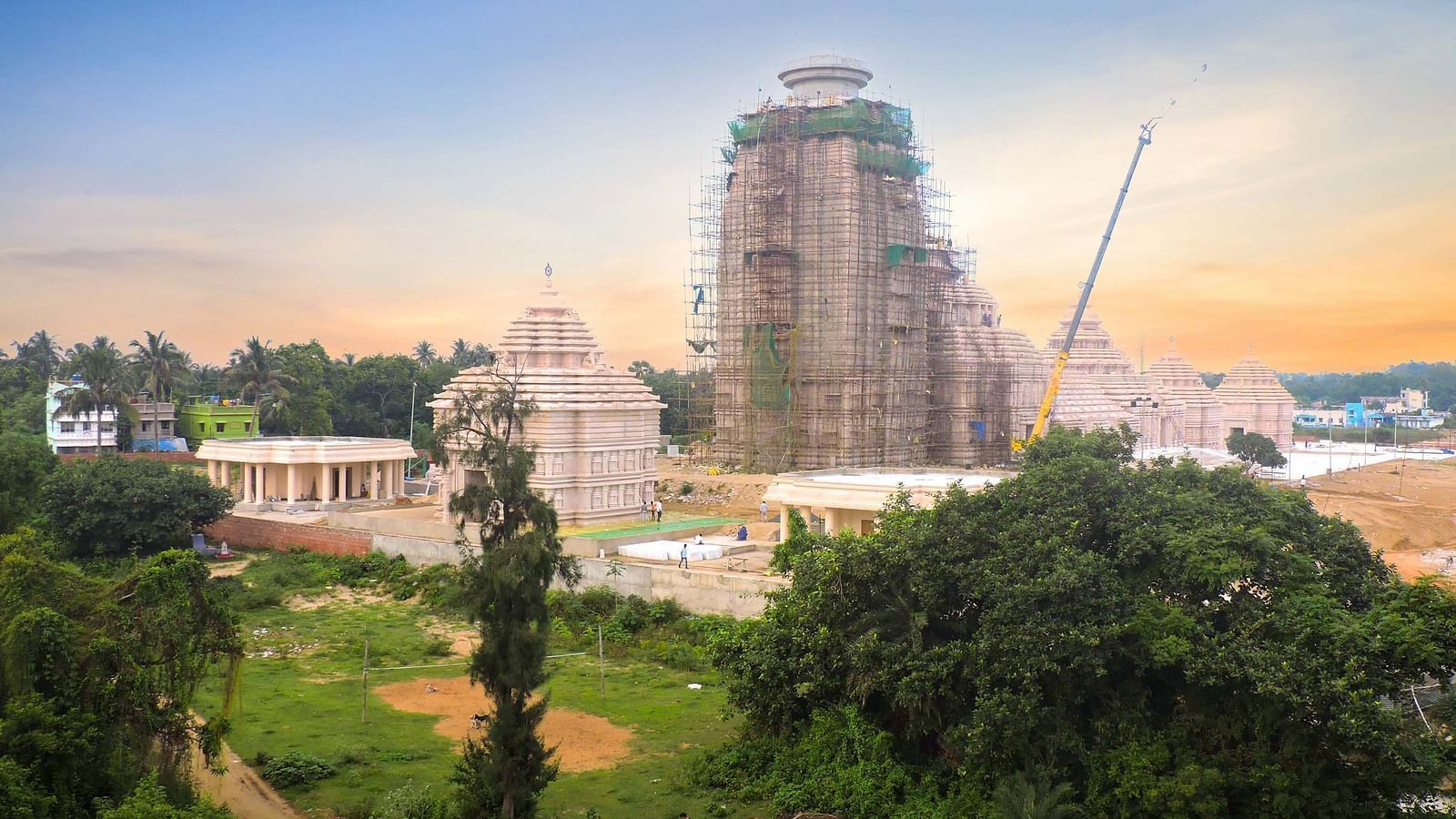 View of Jagannath Temple under construction surrounded by greenery, with scaffolding and cranes in the background, captured during sunset