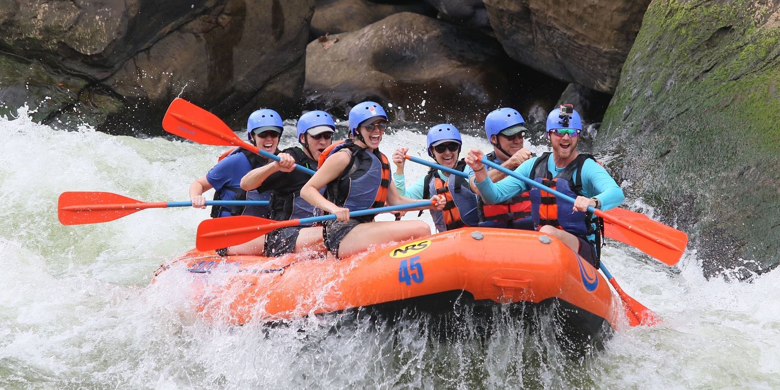 A group of people wearing helmets and life jackets are white-water rafting in a turbulent river.