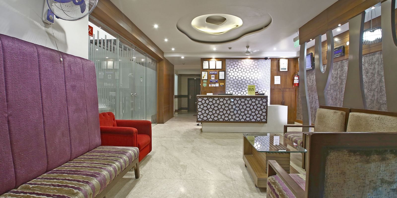 Seating area with modern ceiling design, bright lighting, a brown sofa on the right and a seating space on the left along with a red single chair and a reception desk in the far - Hotel Emerald, Chandigarh