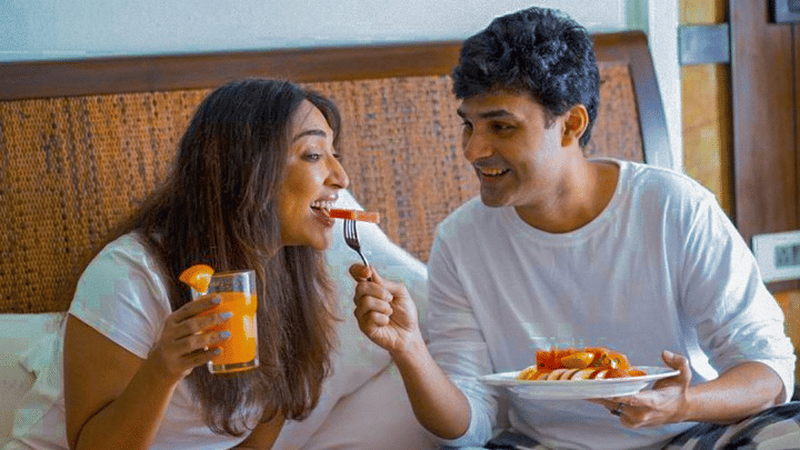 A man and a woman sitting on a bed at Rhythm Lonavala while eating food from the trays placed in front of them as they look at the meal and interact with each other.