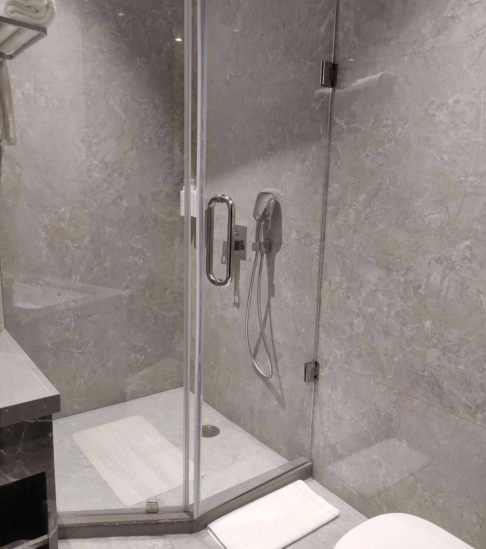 A bathroom at Anant Mahal, Jaipur featuring a glass shower enclosure, grey tiled walls, and white towels on a metal rack.