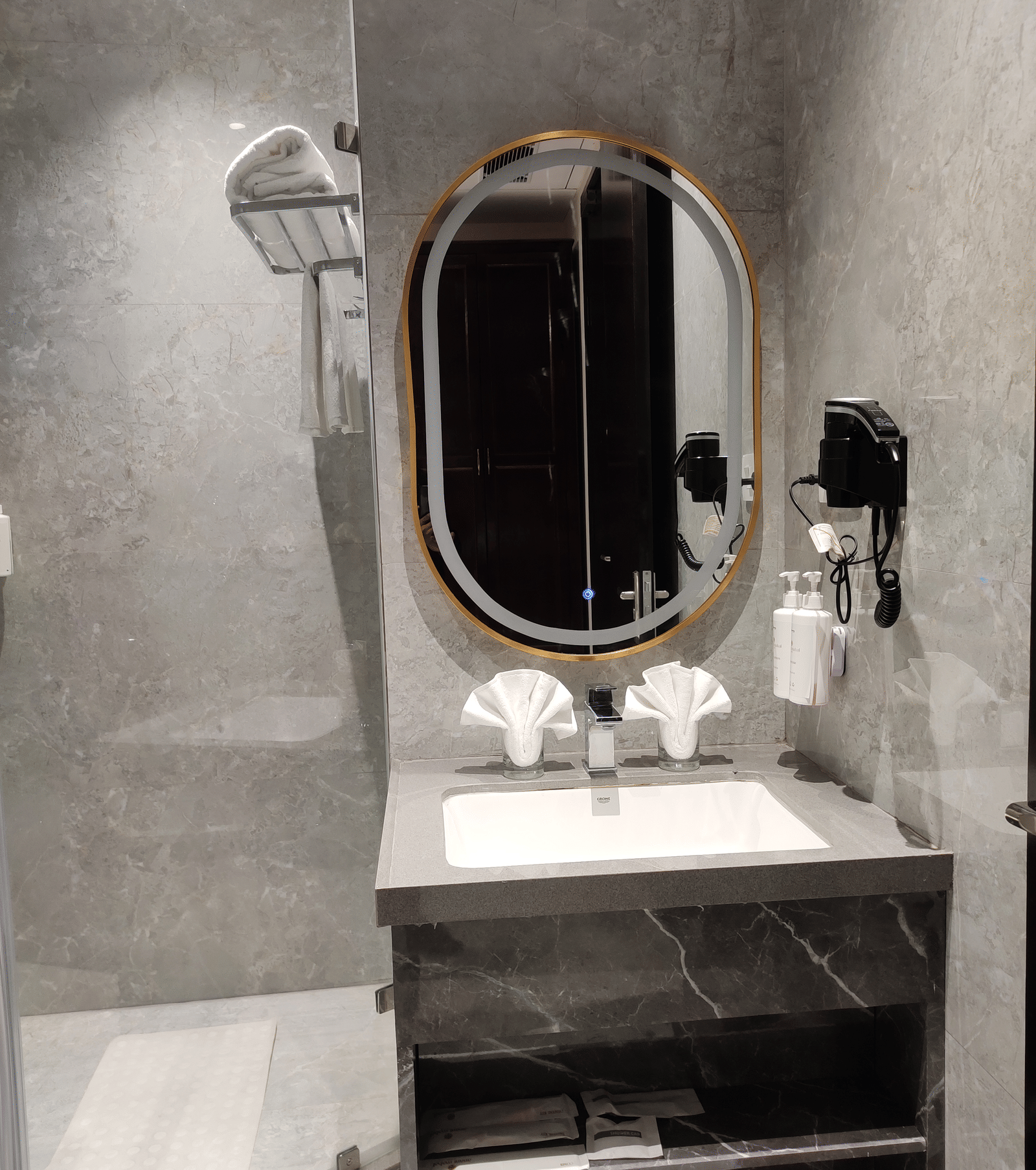 A bathroom at Anant Mahal, Jaipur with a stone vanity, an oval mirror, a wall mounted hair dryer, and a glass shower door.