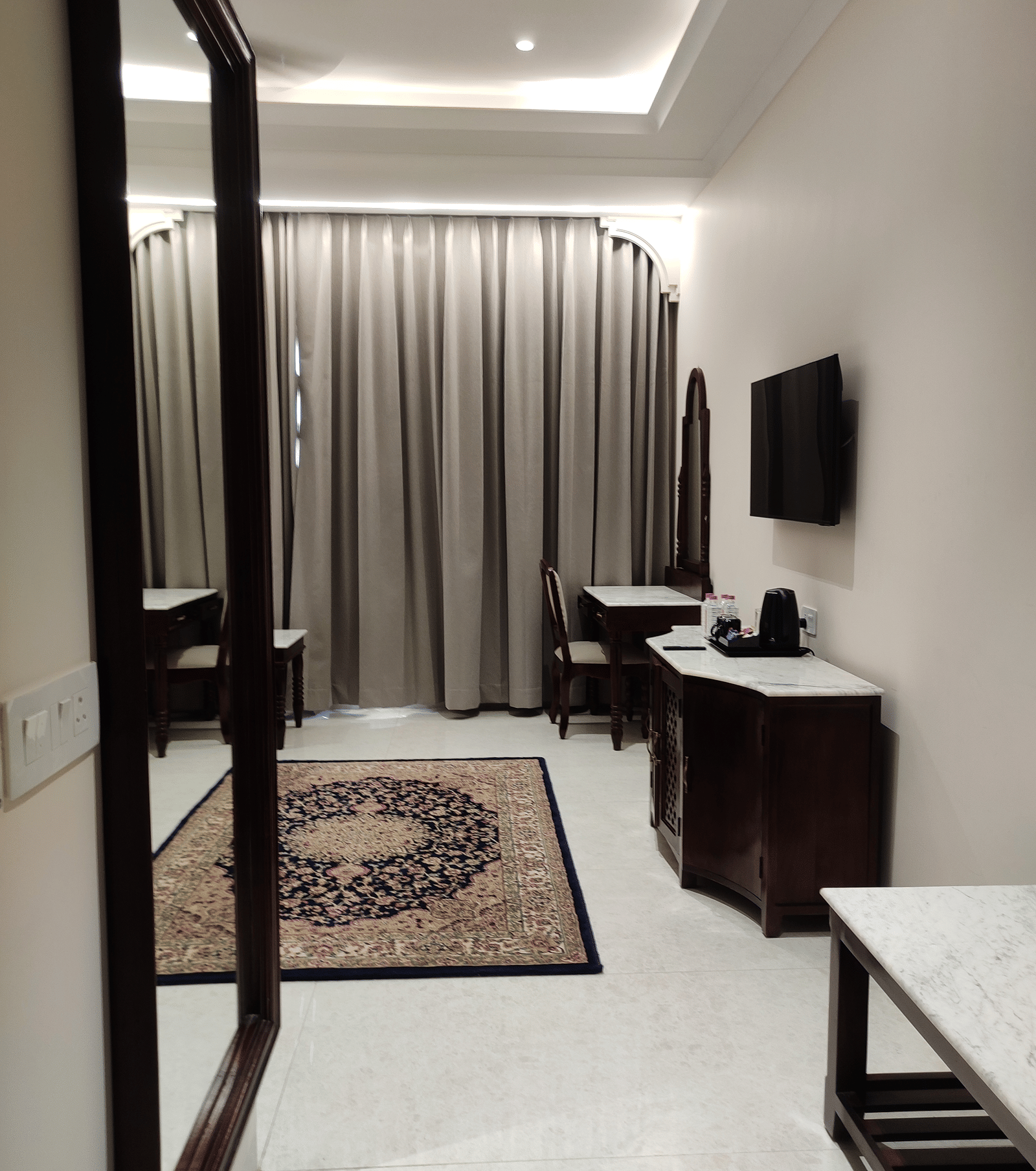 A room at Anant Mahal, Jaipur featuring a large mirror, a patterned rug on the floor, a large window covered by blinds, and a wall mounted television.