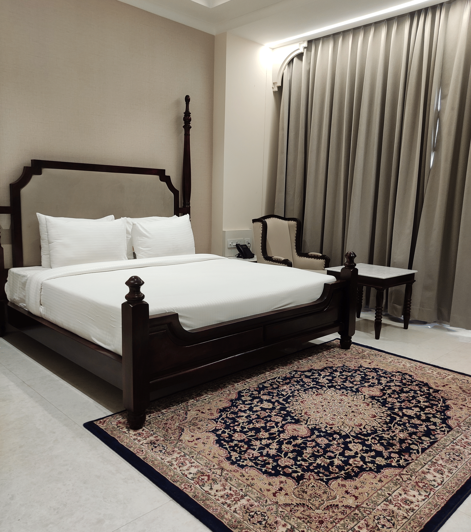 A bedroom at Anant Mahal, Jaipur featuring a king size bed, a patterned rug, floor to ceiling curtains behind a table, and a chair.