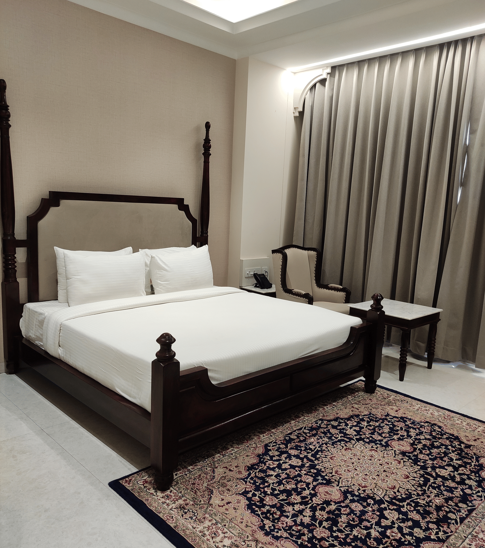 A bedroom at Anant Mahal, Jaipur featuring a king size bed, a patterned rug, floor to ceiling curtains behind a table, and a chair.