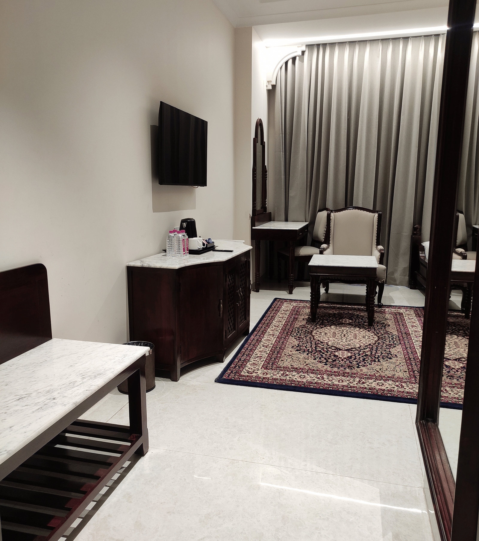 A room at Anant Mahal, Jaipur with a wooden cabinet, a wall mounted television, a patterned rug on the floor, a table and a chair.