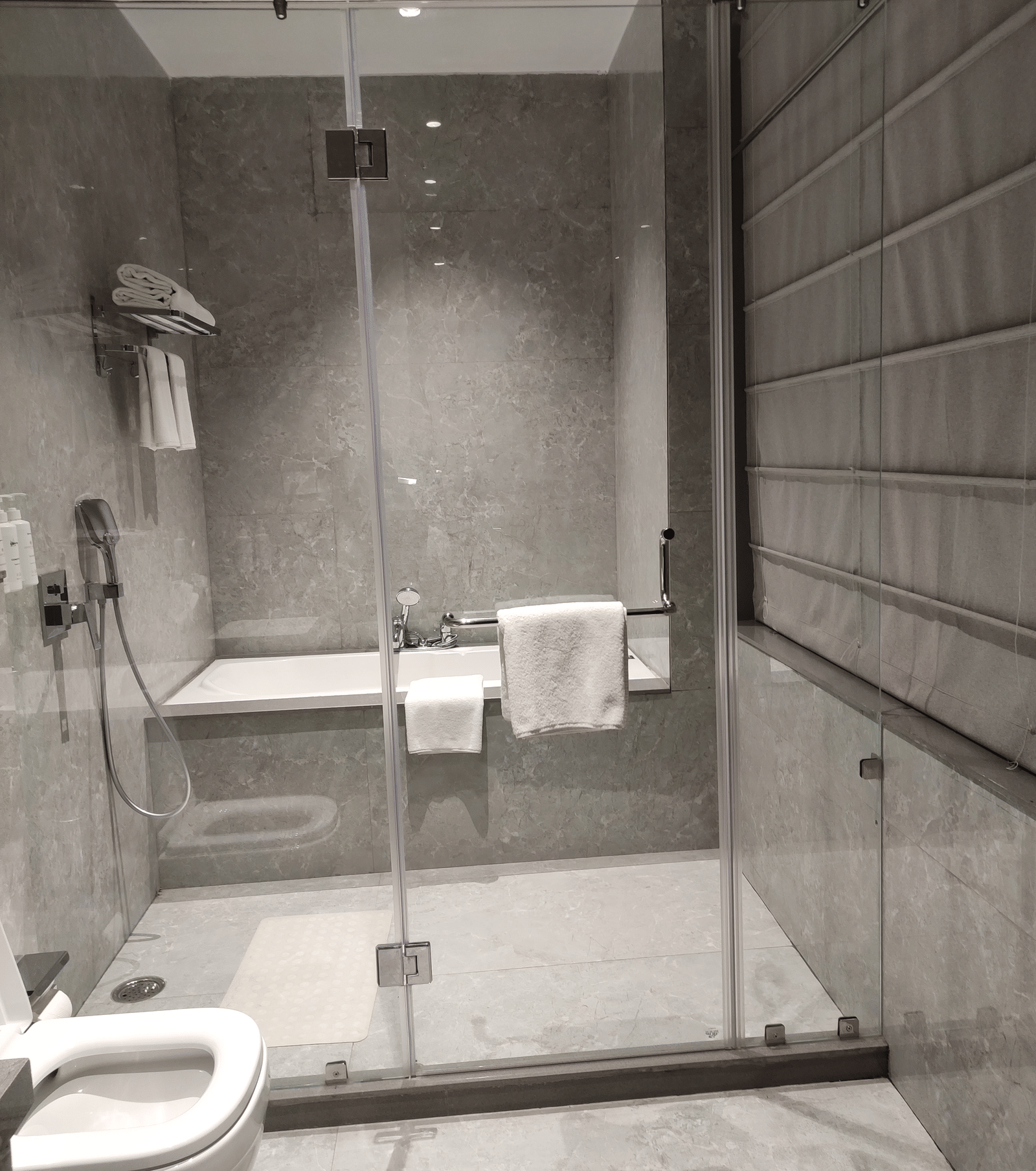 A grey tiled bathroom at Anant Mahal, Jaipur featuring a glass shower door, a white toilet, and 2 white towels hanging on a metal rail.