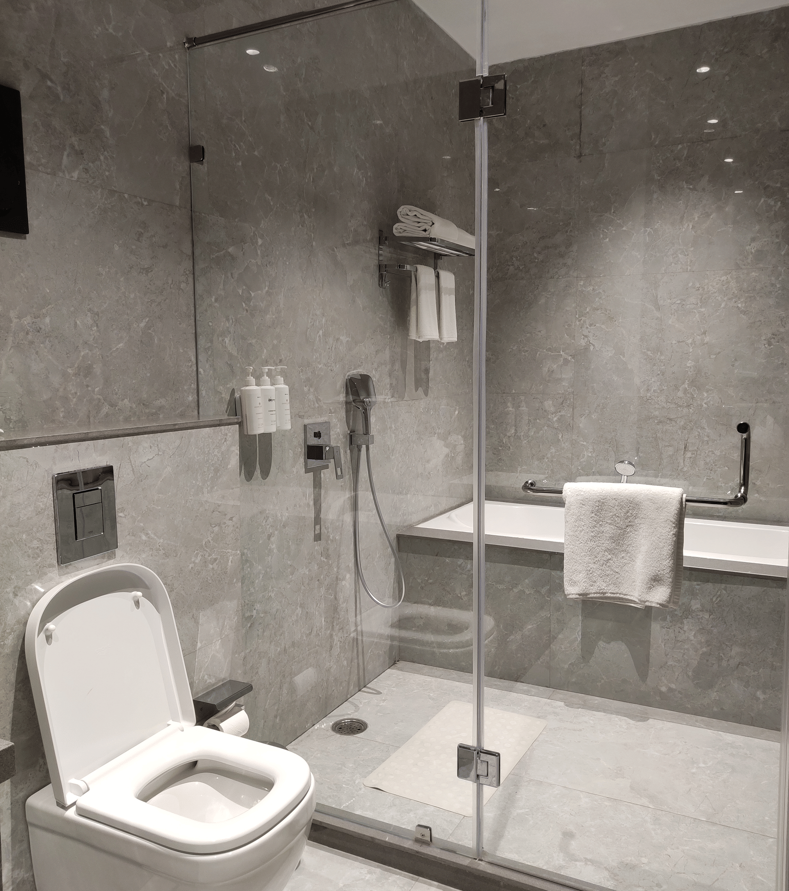 A bathroom at Anant Mahal, Jaipur featuring a glass walk in shower, a white toilet, and grey tiled walls with a chrome towel rack.