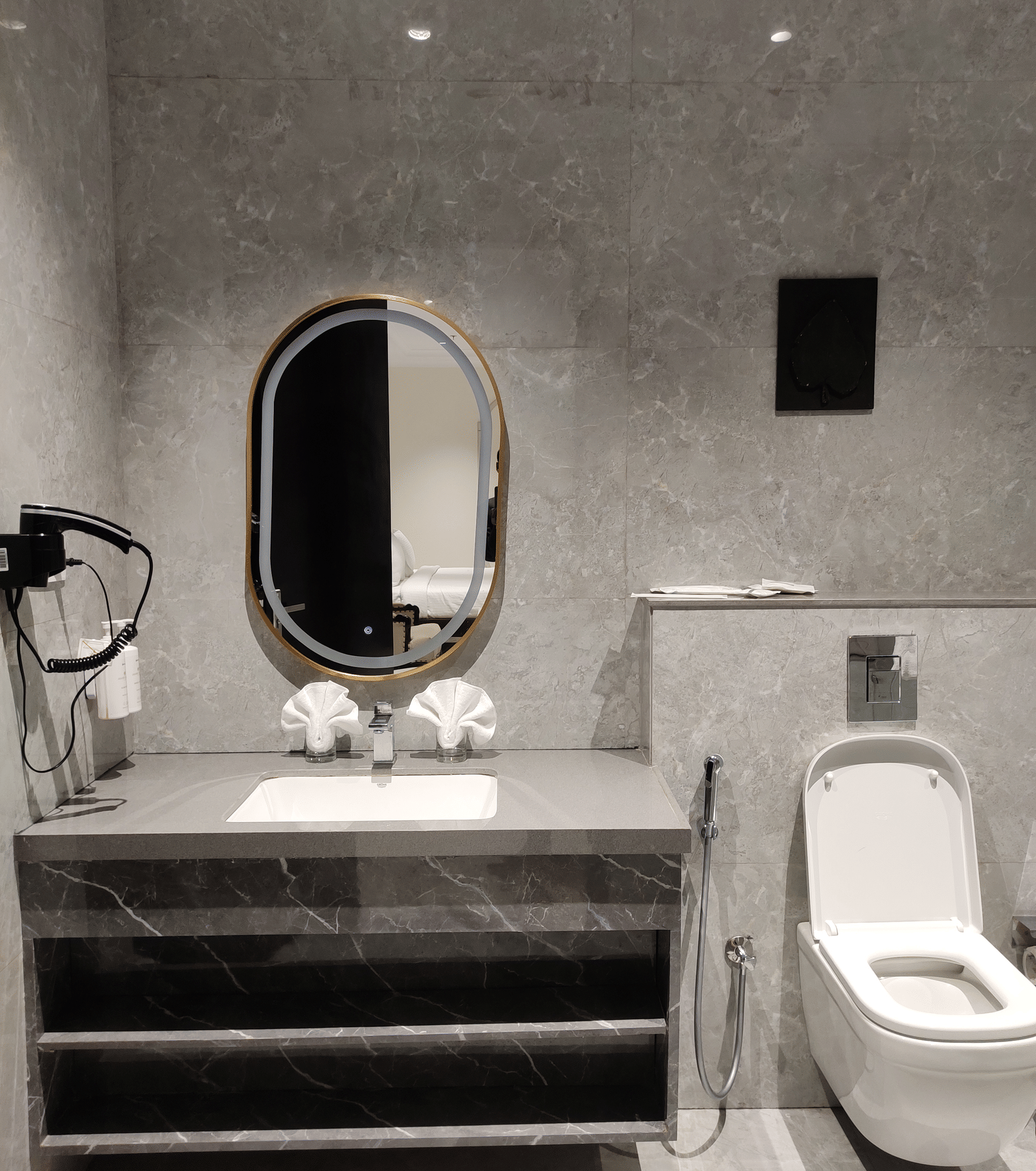 Bathroom interior at Anant Mahal, Jaipur with a stone vanity, an oval mirror, a wall mounted hair dryer, and a white toilet.