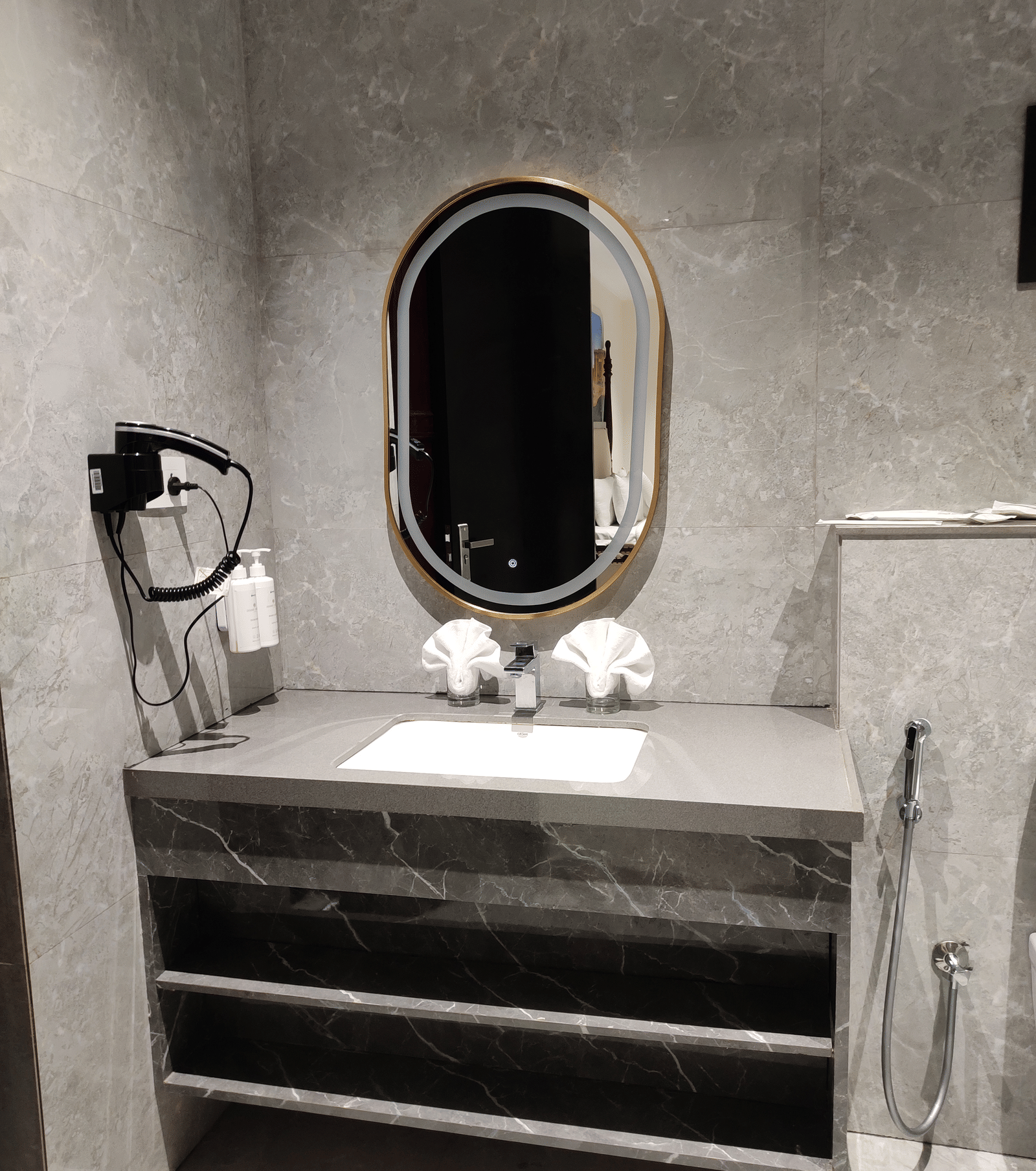 A bathroom at Anant Mahal, Jaipur featuring a stone countertop with a sink, open wooden shelving, a hair dryer, and an oval mirror.