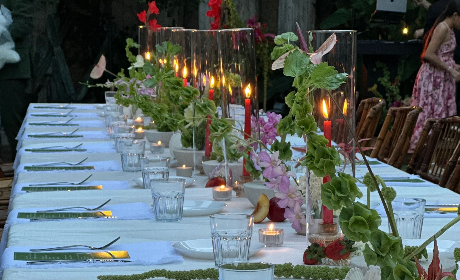 A long dinner table where several items are placed including forks, menu cards, glass,  etc. The table is also decorated with candles wrapped in a glass and artificial flower pots with flowers and leaves.