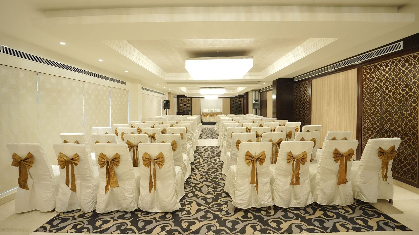 A banquet hall with rows of chairs draped in white covers and gold bows, ready for an event - Fragrant Nature Cochin