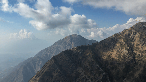 A panoramic view of dark, rugged mountain peaks stretching toward a blue sky with white clouds at Ananta Express, Katra.
