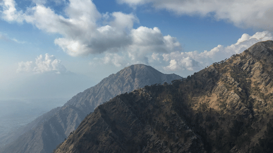 A panoramic view of dark, rugged mountain peaks stretching toward a blue sky with white clouds at Ananta Express, Katra.