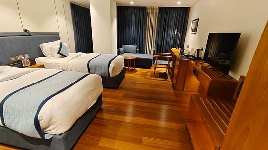 Deluxe Twin Room at Pride Hotel Udaipur featuring a couple of twin bed facing the TV mounted on the wall and a sitting space made closer to the window covered with curtains