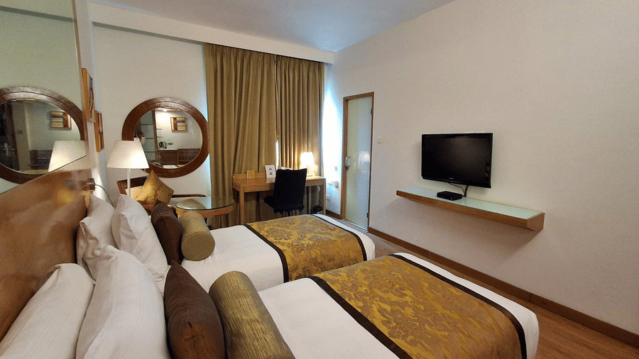 An angled view showing the two single beds in the Deluxe Twin Room at Pride Premier Chennai. A television is mounted on the wall opposite the beds, and curtains cover the window at the back.