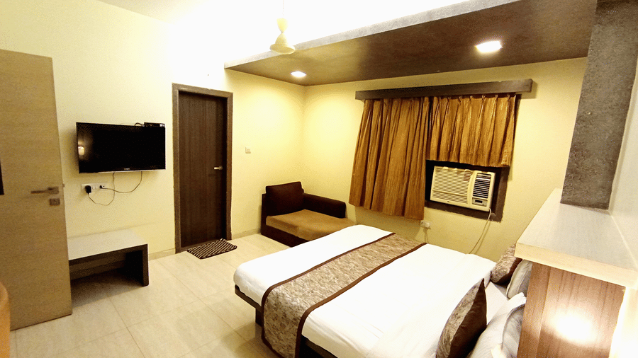 Well-furnished executive room showing a double bed, wall-mounted television, window air conditioner, and a small seating stool.