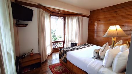 A double bed facing a curtained glass window in the Executive Suite of Cedar Inn in Darjeeling that has wooden flooring, a TV.