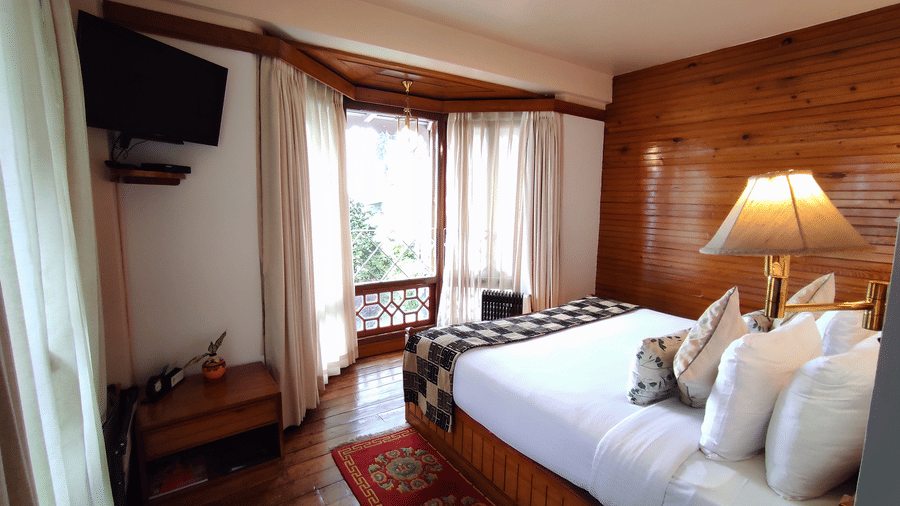 Bedroom of the Executive Suite at Cedar Inn featuring a large bed with cushions, bedside lamps, wooden interiors, a wall mounted television, and windows allowing natural light.