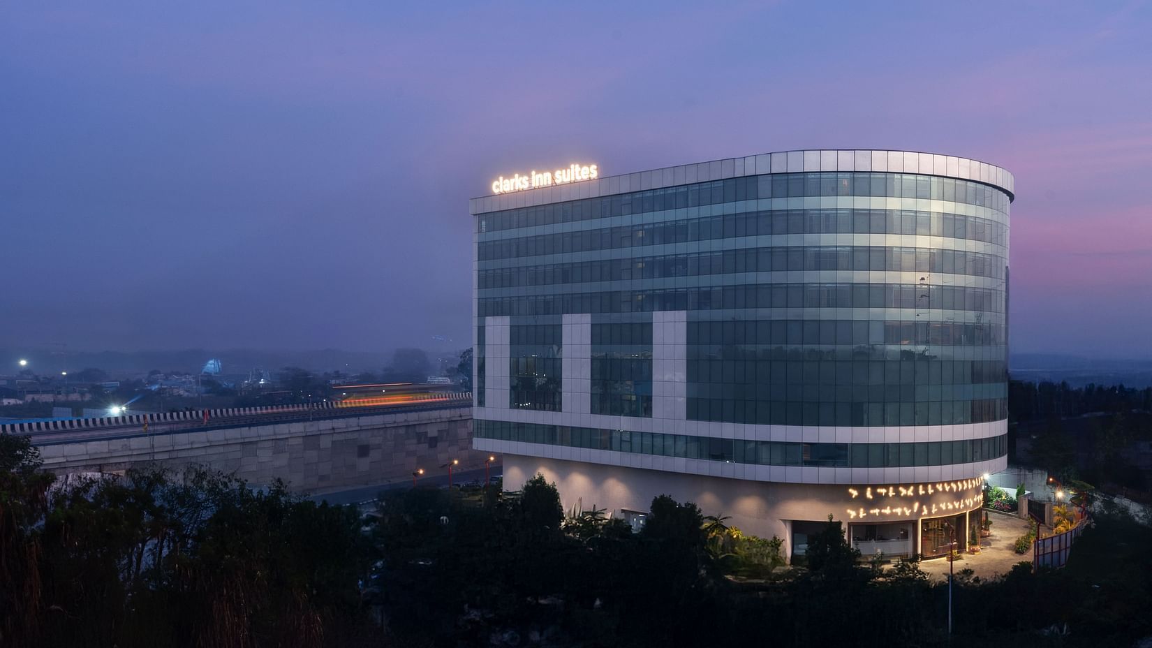 Evening shot of a curved glass hotel building with illuminated signage against a dusky sky at Clarks Inn Suites, Hyderabad