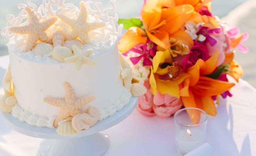 A wedding cake decorated with starfish sits next to a vibrant orange and pink bouquet on a white table at Casa Morada.