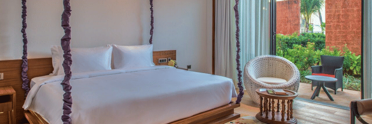 The Deluxe Patio King Room with a large wooden bed hanging from the ceiling on purple rope vines, and an armchair at Azaya Beach Resort, Goa.