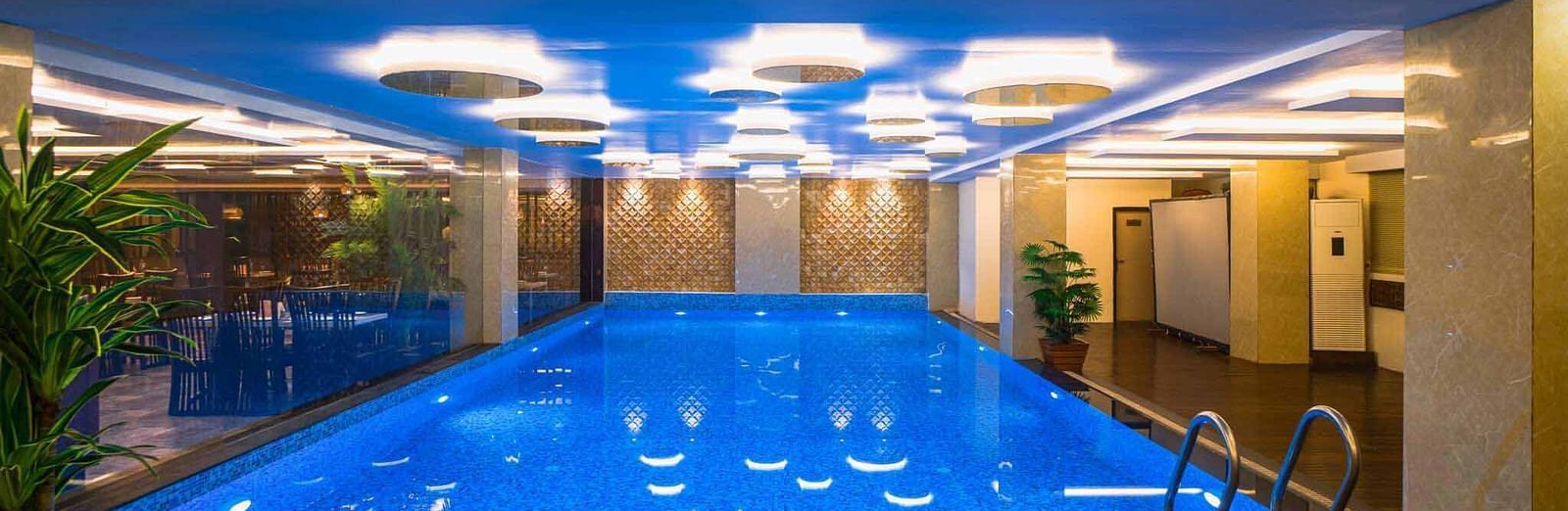 An indoor swimming pool at Mansha Hotel Regency with blue water and a light blue ceiling, surrounded by pillars and lit by overhead lights.