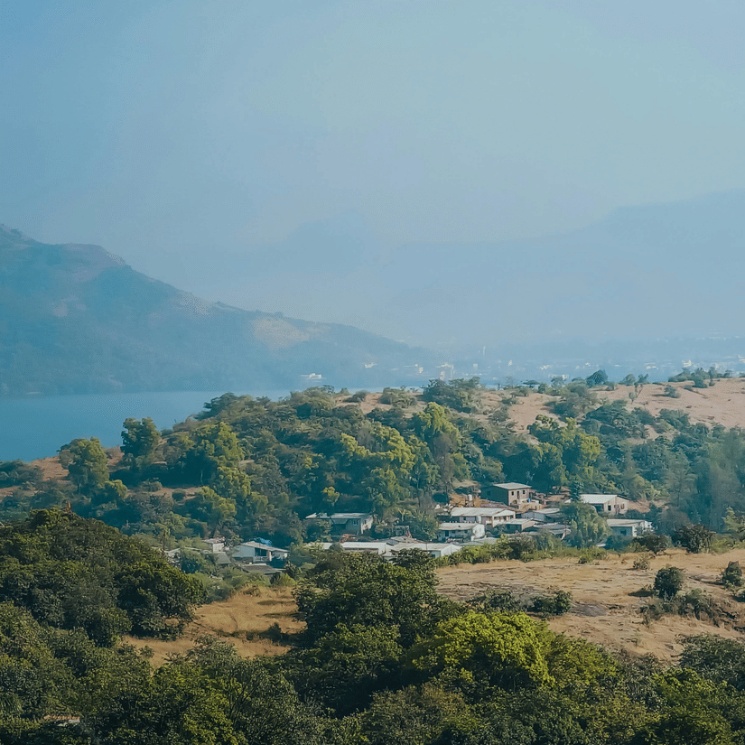 An aerial view of Lonavala with a lake and mountains in the distance, and buildings covered by trees in front of it.