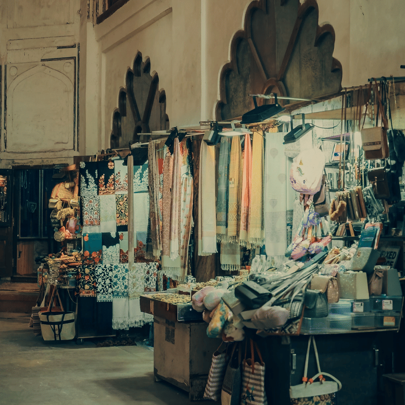 Colourful textiles and garments hang on display inside a rustic market stall with traditional arches.