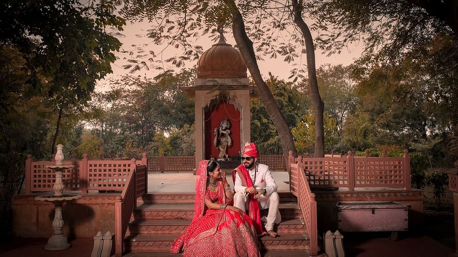 A newlywed couple in wedding attire sits on temple stairs, sharing a joyful moment in a serene outdoor setting.- Umaid Palace - A Lakeside Heritage Palace Resort