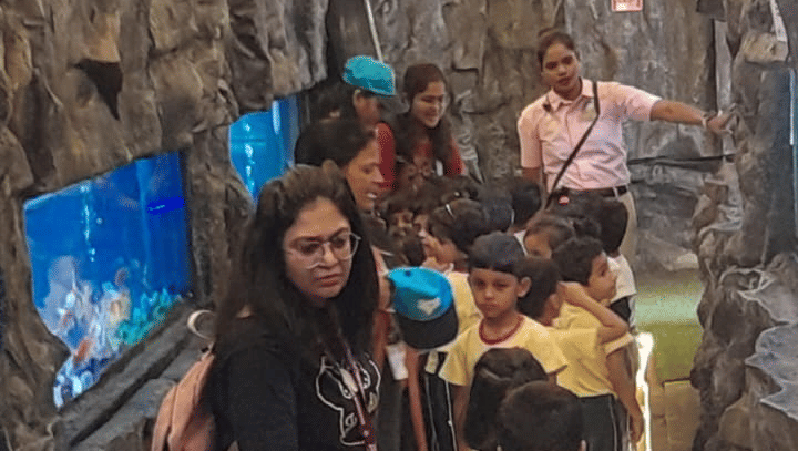A vertical, brightly lit interior view of a crowded area at EsselWorld Fish Paradise, possibly a tunnel or hallway inside the aquarium, filled with many visitors.